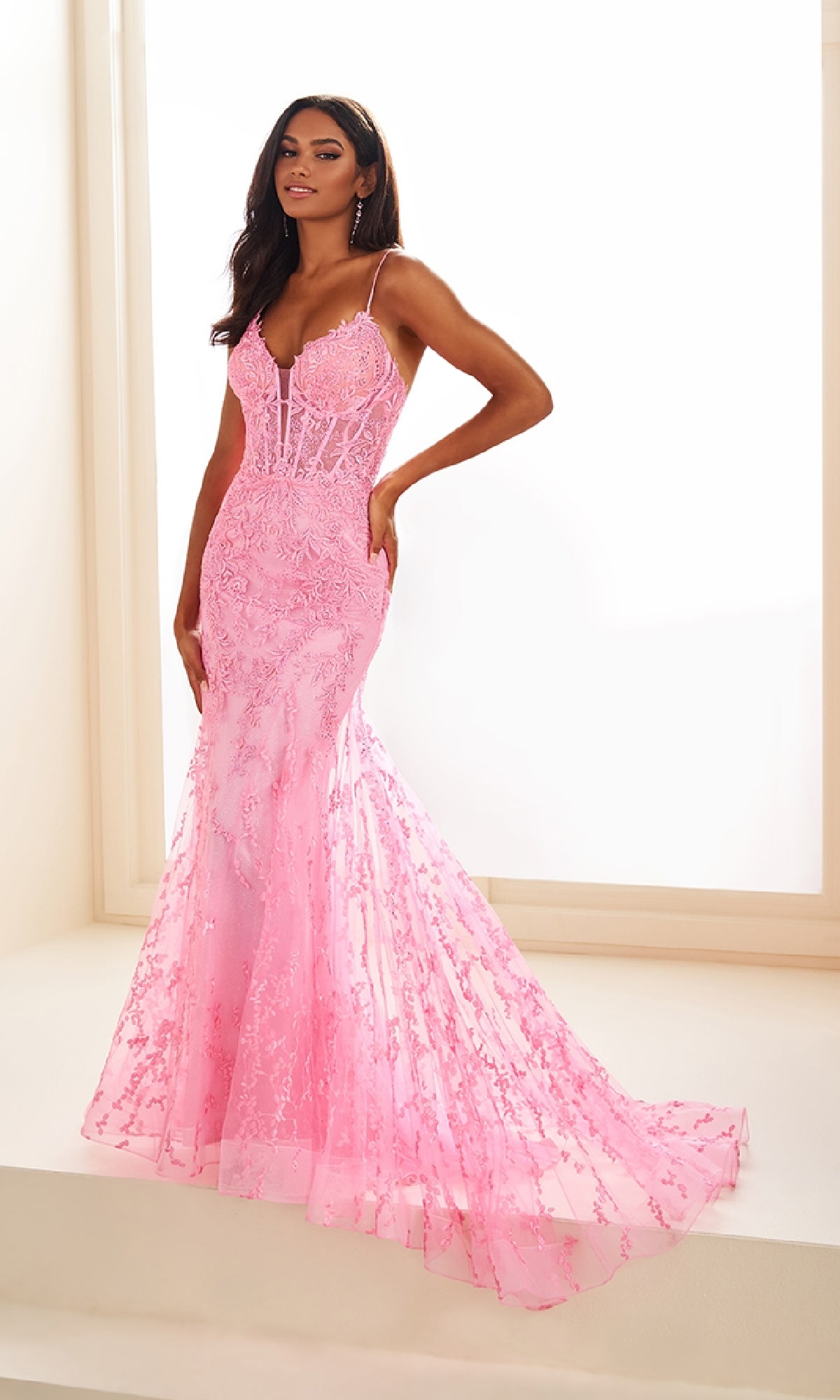 A woman stands in a bright room wearing the Ellie Wilde Beaded-Tulle Long Prom Dress EW35010—a fitted, floor-length pink gown with thin straps, a flared skirt, and delicate beaded embroidery.